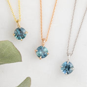 Three necklaces with round blue gemstones set in gold-colored metal, displayed on a white background with a green leaf to the side.