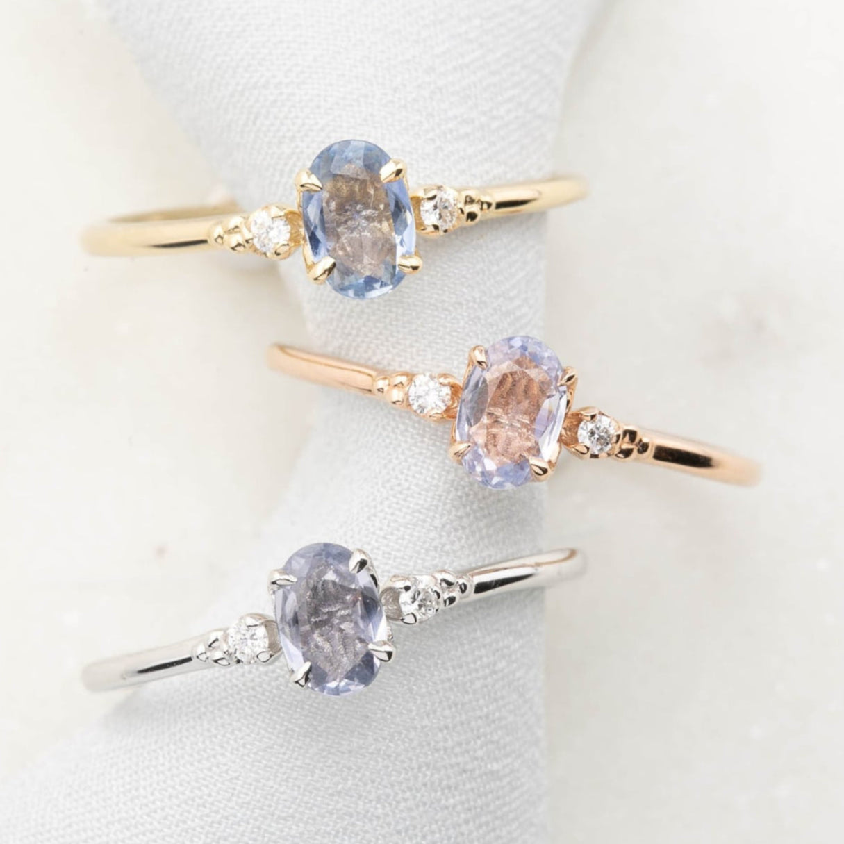 Three 'Estel Rings' with oval-shaped sapphires, in settings of 14k rose gold, 14k white gold, and 14k yellow gold, displayed against a light background., oval shape gemstone