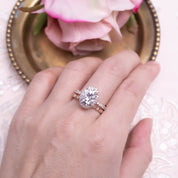 Close-up of hand wearing a 3ct Neo Moissanite halo ring with rose gold half-eternity band and pink flower background