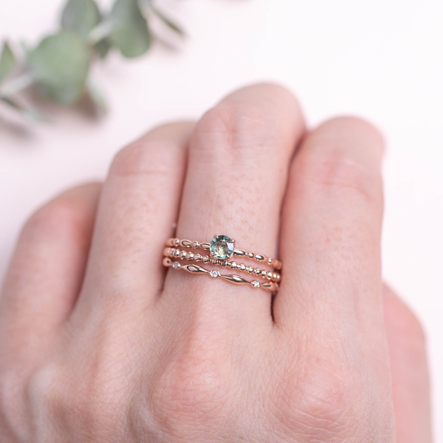 Hand wearing three delicate rose gold rings with textured bands and a small green gemstone centerpiece