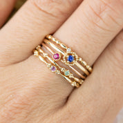 Stack of delicate 14k gold birthstone rings with gemstones in pink, blue, and green on a finger.