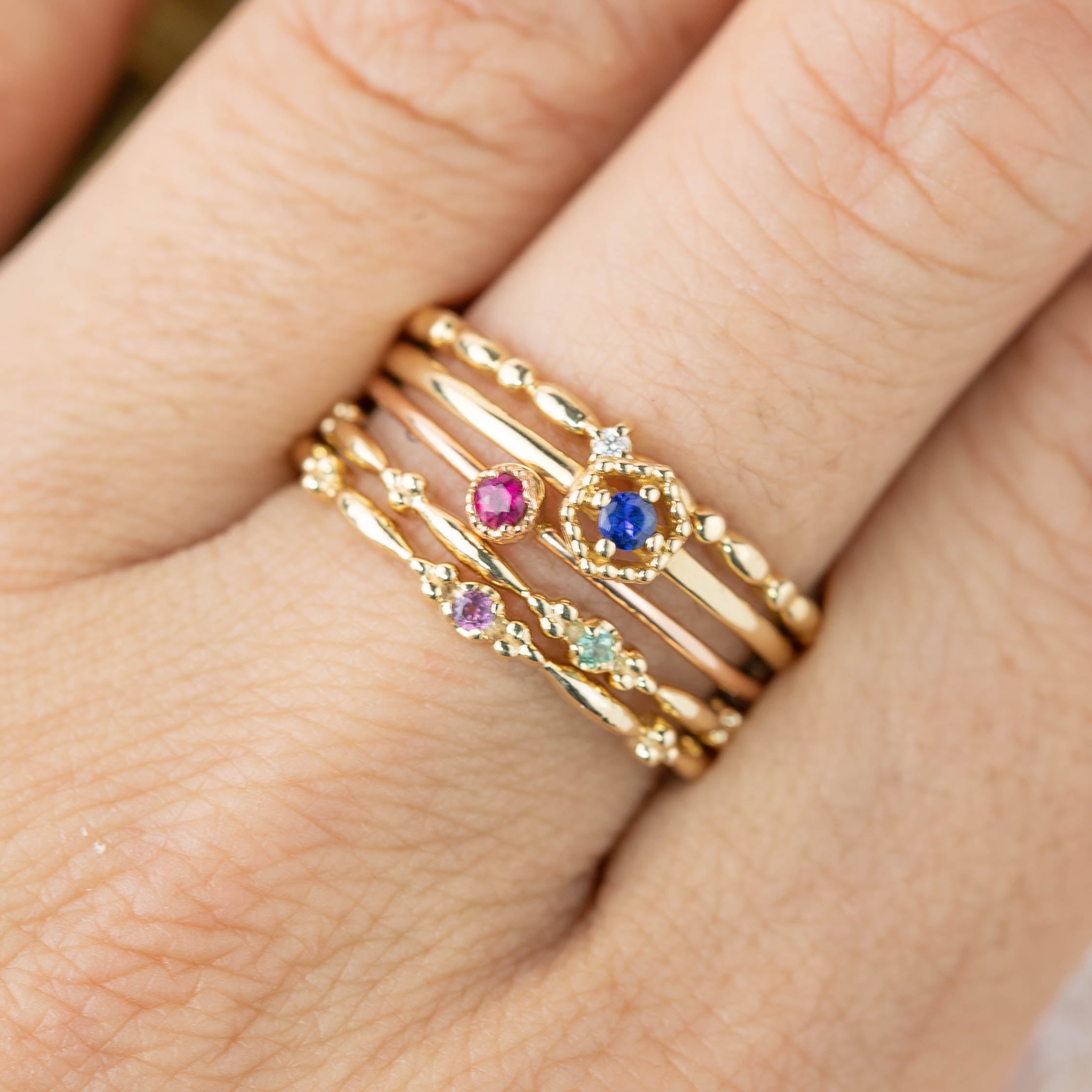 Stack of delicate 14k gold birthstone rings with gemstones in pink, blue, and green on a finger.