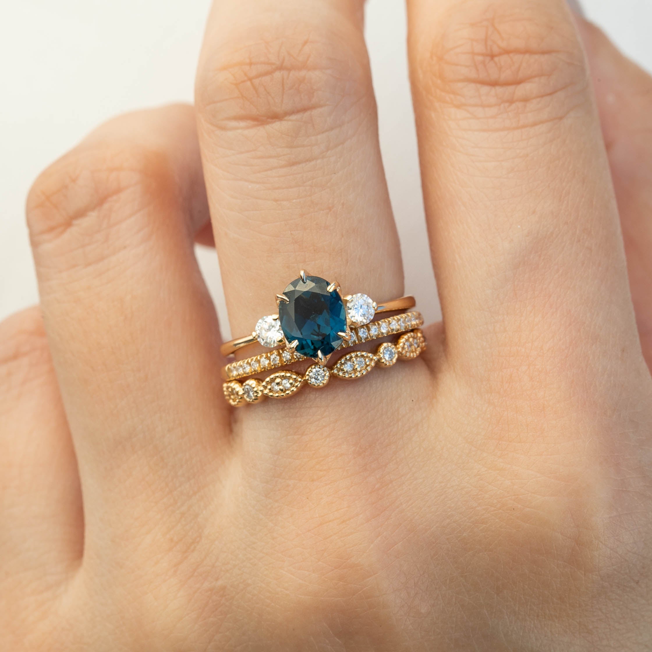 Hand wearing three gold rings including a gold band with diamonds and a ring with a large dark blue gemstone centerpiece