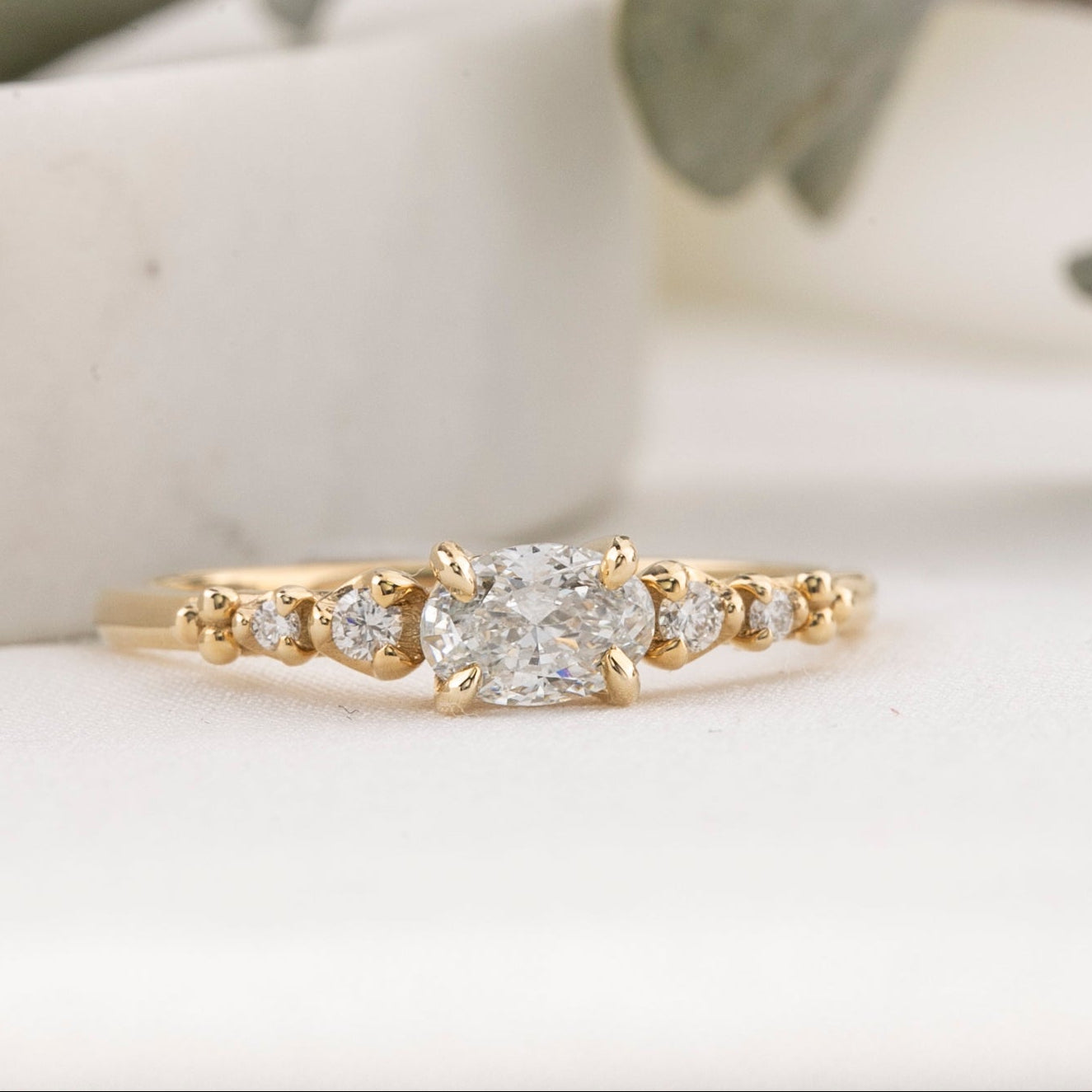Gold ring with a diamond on a white surface with green leaves in the background