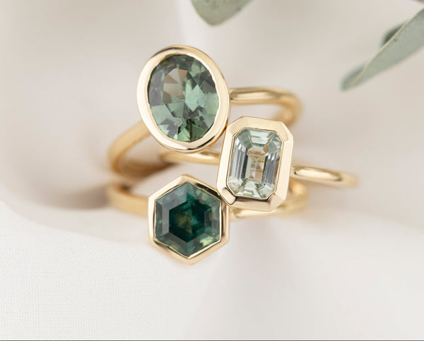 Three gold rings with green gemstones on a light background with eucalyptus leaves.