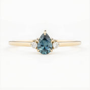 A pear-cut teal Montana sapphire ring with accent diamonds set in a 14k yellow gold band, displayed against a white background.