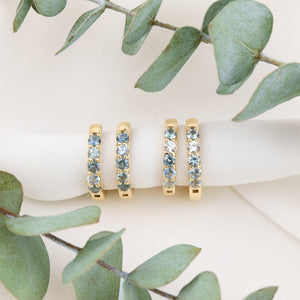 Three gold eternity rings with light blue gemstones are displayed on a white ribbon, framed by green eucalyptus leaves on a light beige background.