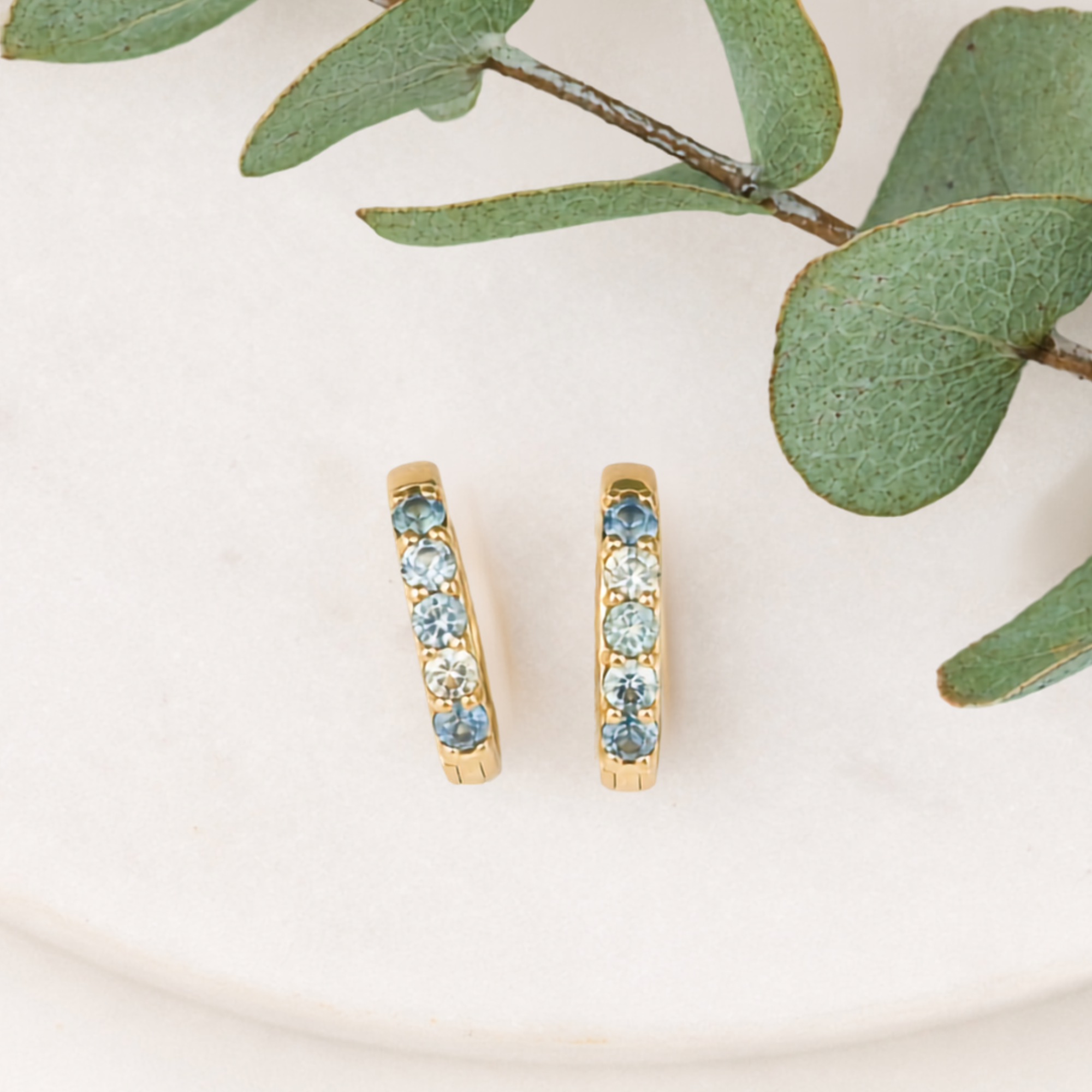 A pair of gold huggie hoop earrings with a row of light blue round gemstones, displayed on a light beige textured surface with green eucalyptus leaves.