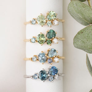 Set of gold rings with green and blue gemstones on a white stand with a plant in the background.