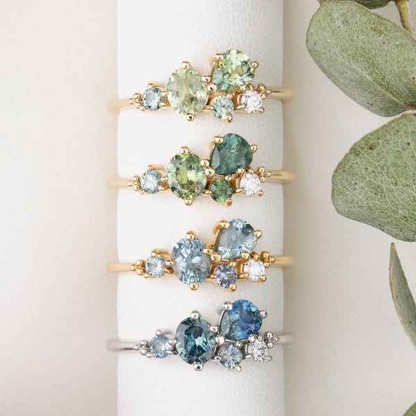 Set of gold rings with green and blue gemstones on a white stand with a plant in the background.
