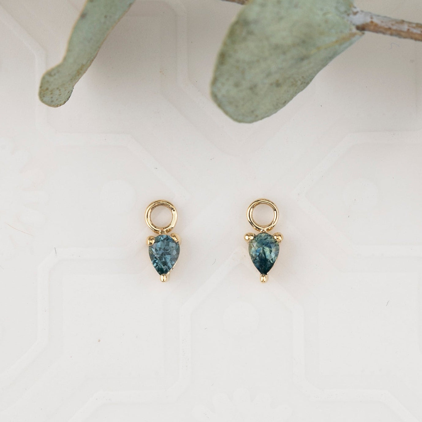 Gold earrings with blue teardrop stones on a white background with green leaves.