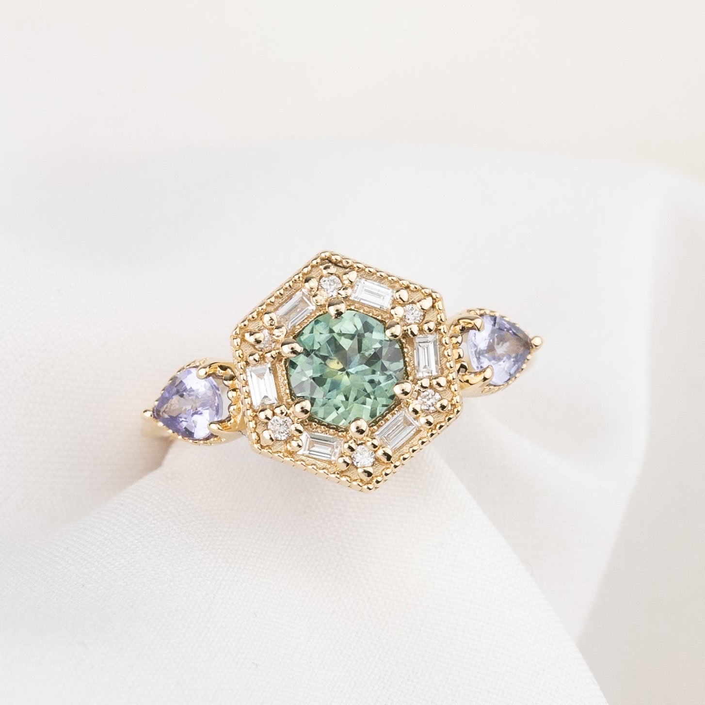 14k hexagonal gold ring with a central green round green Montana gemstone, surrounded by baguette and round accent diamonds and flanked by two pear-cut light purple gemstones. 