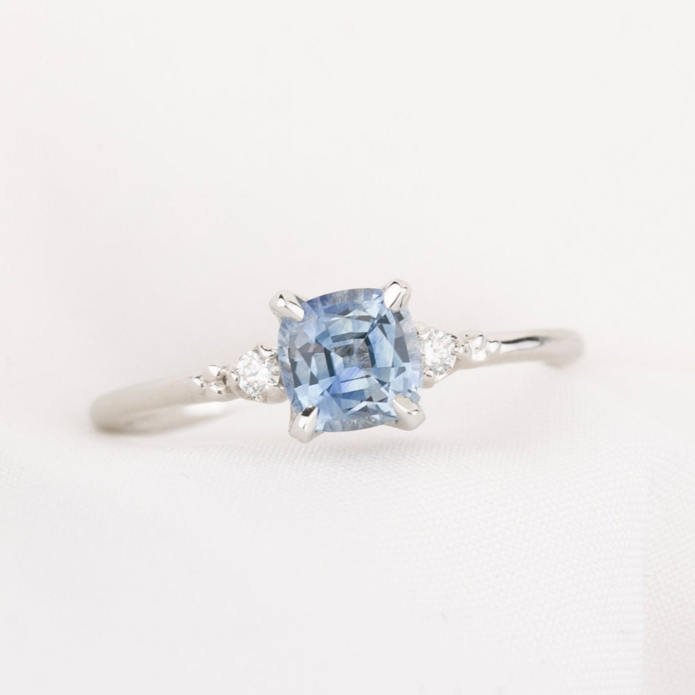 white gold ring with blue light blue cushion cut montana sapphire, with two natural accent diamonds on white background