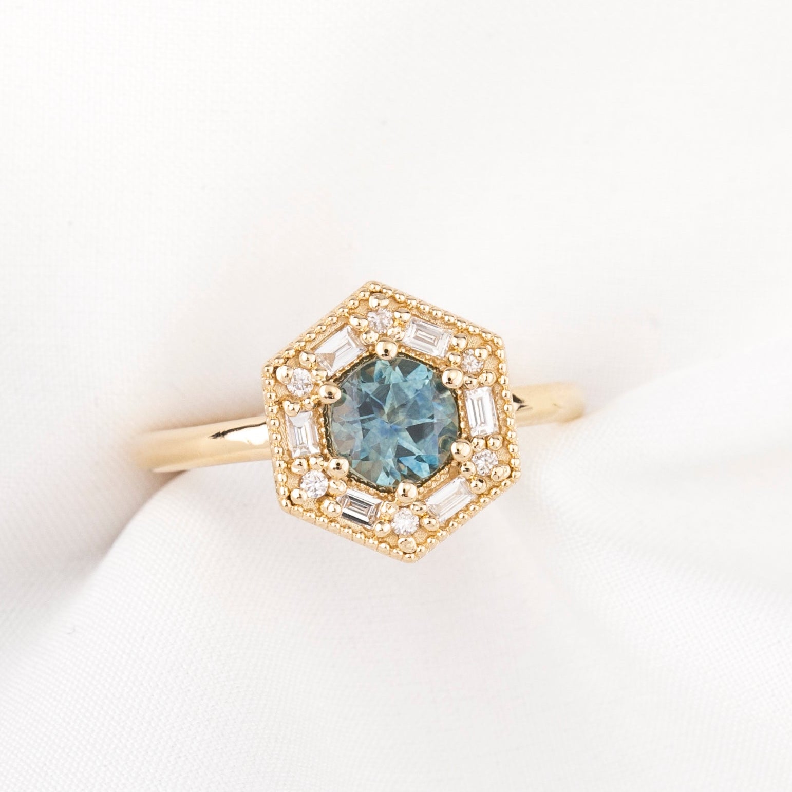 A gold hexagonal halo ring featuring a round blue gemstone in the center, surrounded by small clear stones and baguette accents, displayed on a soft white fabric surface.