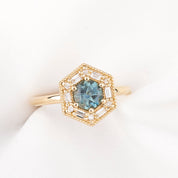 A gold hexagonal halo ring featuring a round blue gemstone in the center, surrounded by small clear stones and baguette accents, displayed on a soft white fabric surface.