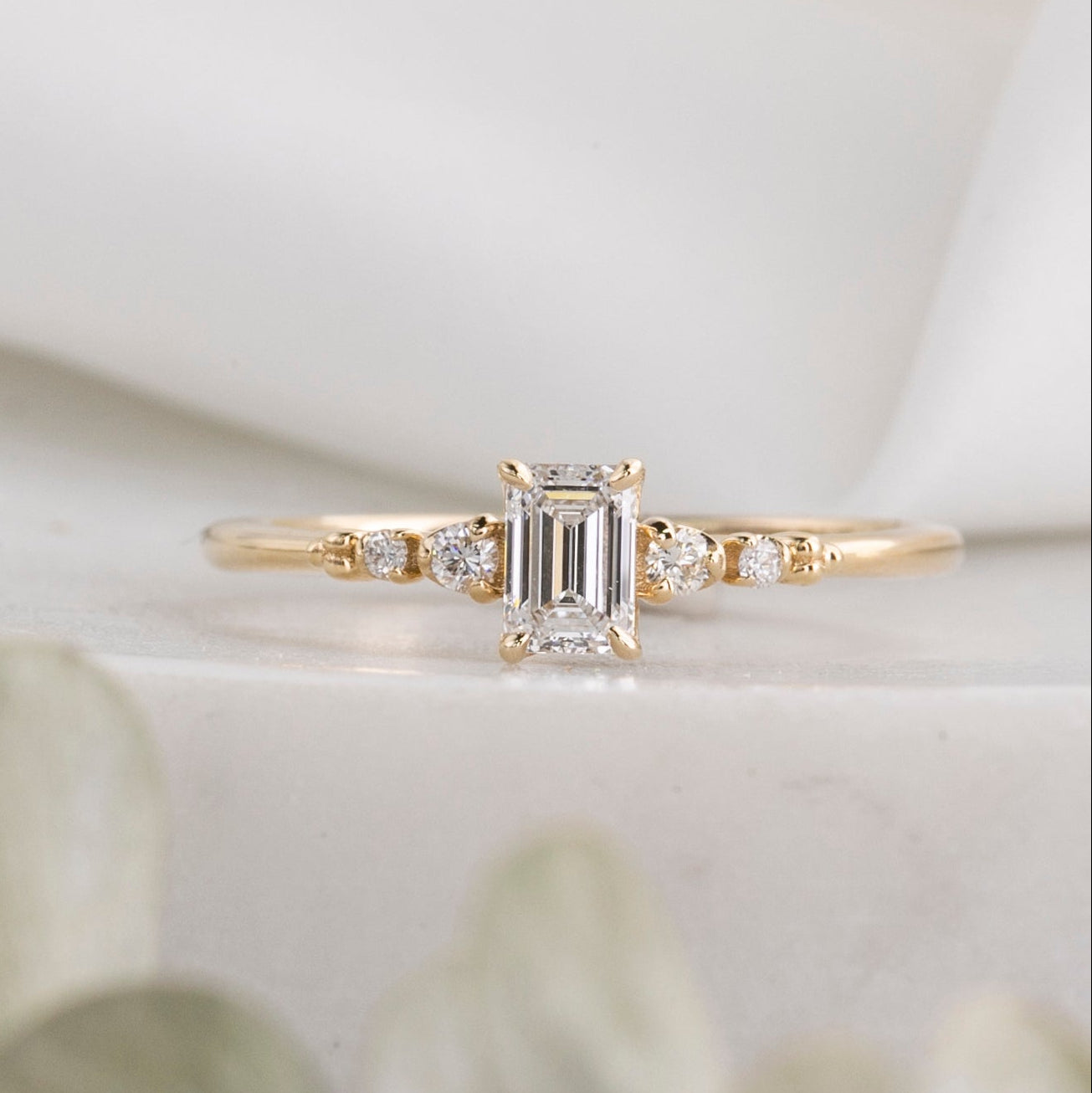 An emerald cut diamond ring with four prongs, set in a tapered band, displayed against a white background with a leafy texture to the side.