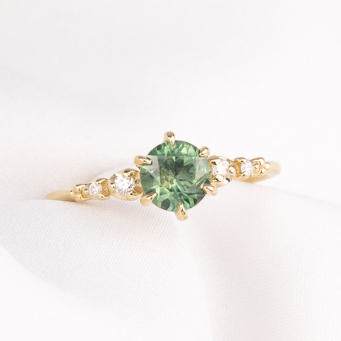 A yellow gold Montana Sapphire ring featuring a round green gemstone in the center and small natural accent diamonds on the band, six prongs engagement ring