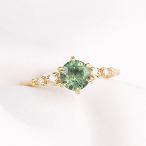 A yellow gold Montana Sapphire ring featuring a round green gemstone in the center and small natural accent diamonds on the band, six prongs engagement ring