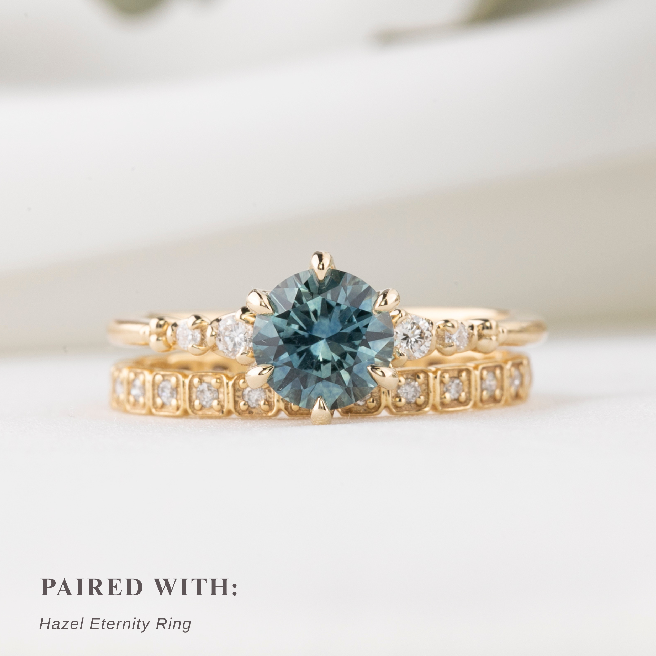 Gold ring with a blue gemstone center stone and natural accent diamonds and matching eternity ring (hazel) on a light background