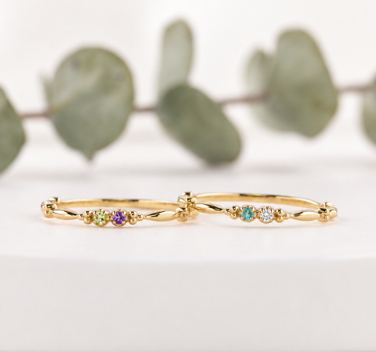 Two dainty 14K yellow gold double birthstone rings with petite round gemstones and delicate beaded details, styled on a soft white background.