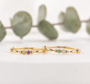 Two dainty 14K yellow gold double birthstone rings with petite round gemstones and delicate beaded details, styled on a soft white background.