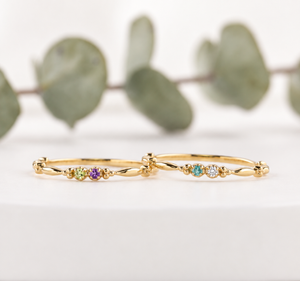 Two dainty 14K yellow gold double birthstone rings with petite round gemstones and delicate beaded details, styled on a soft white background.