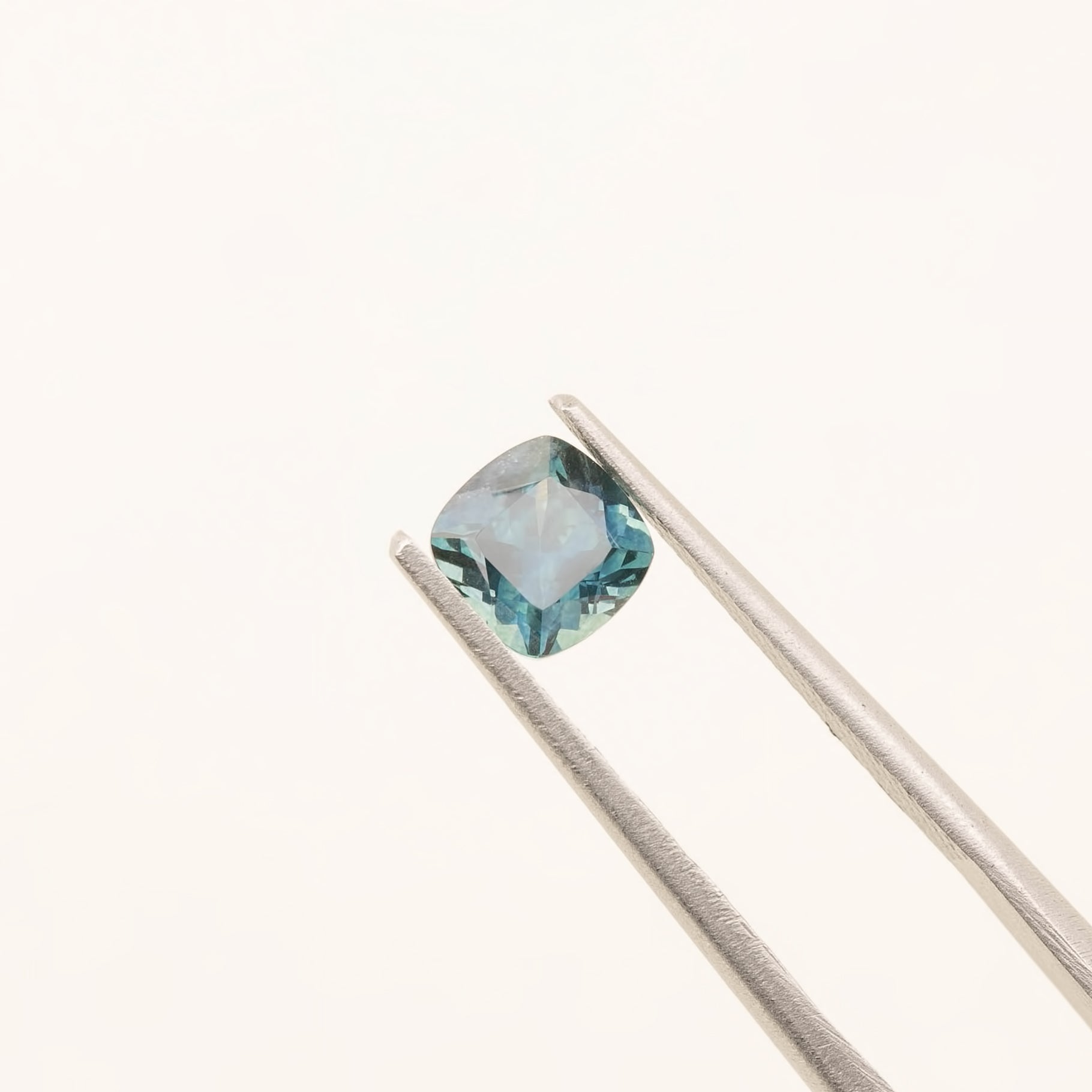 1.42ct cushion cut teal blue Montana sapphire held by tweezers on a light background