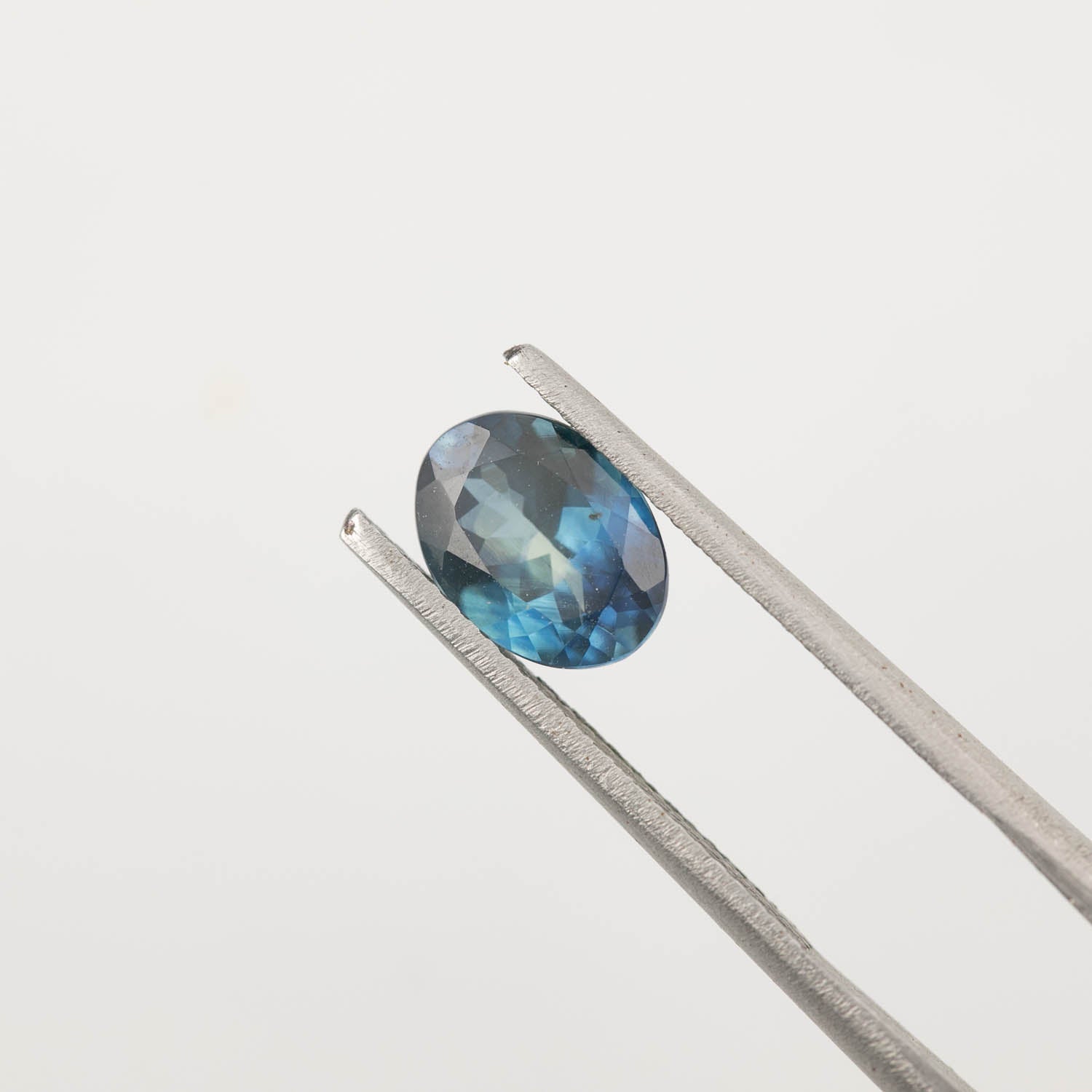1.62ct oval cut teal blue Montana sapphire held by silver tweezers on white background