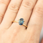 1.62ct oval cut teal blue Montana sapphire ring on finger with gold band, eye clean with small inclusions visible