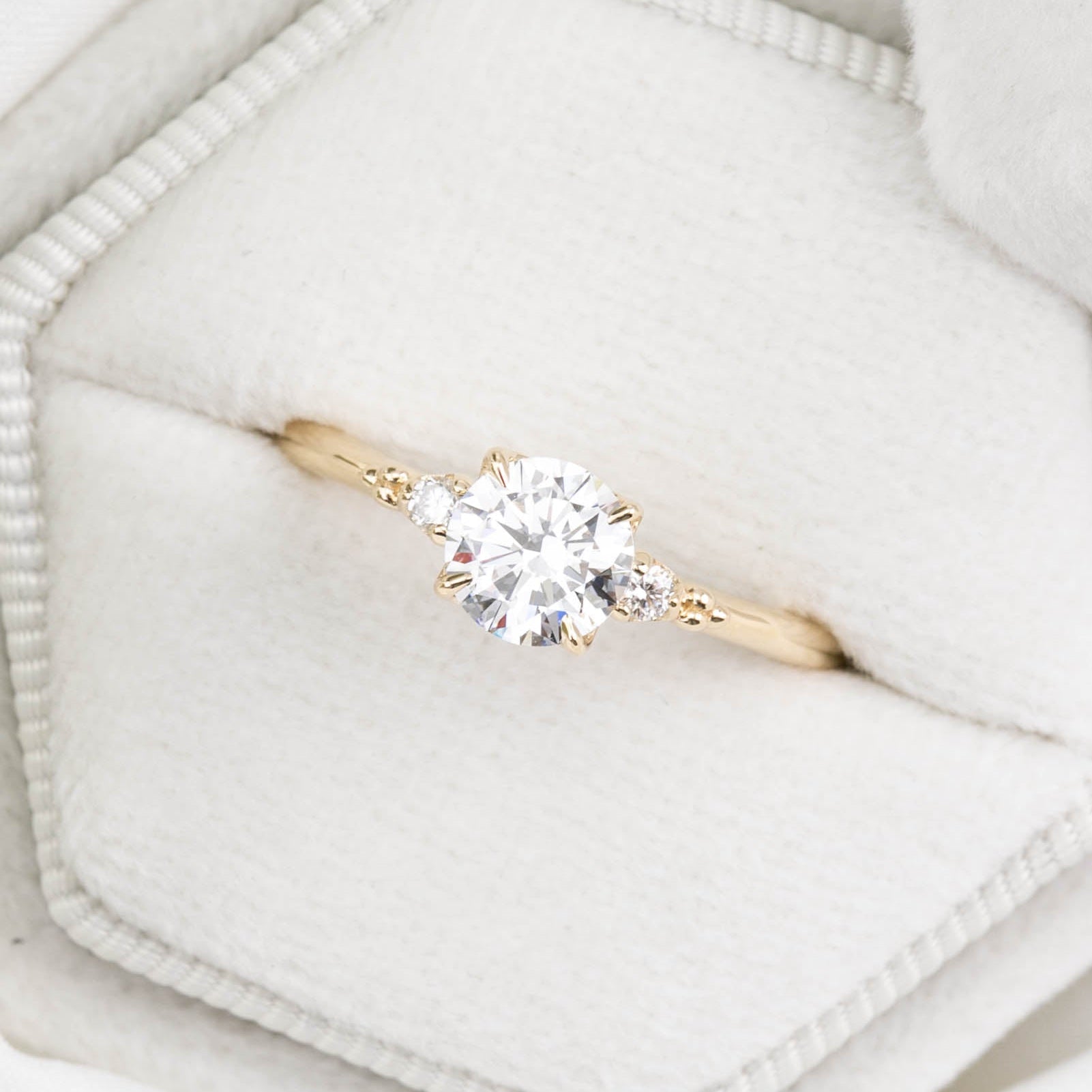 A round lab diamond ring with a yellow gold band, displayed in a white hexagonal box with a green leaf in the background.